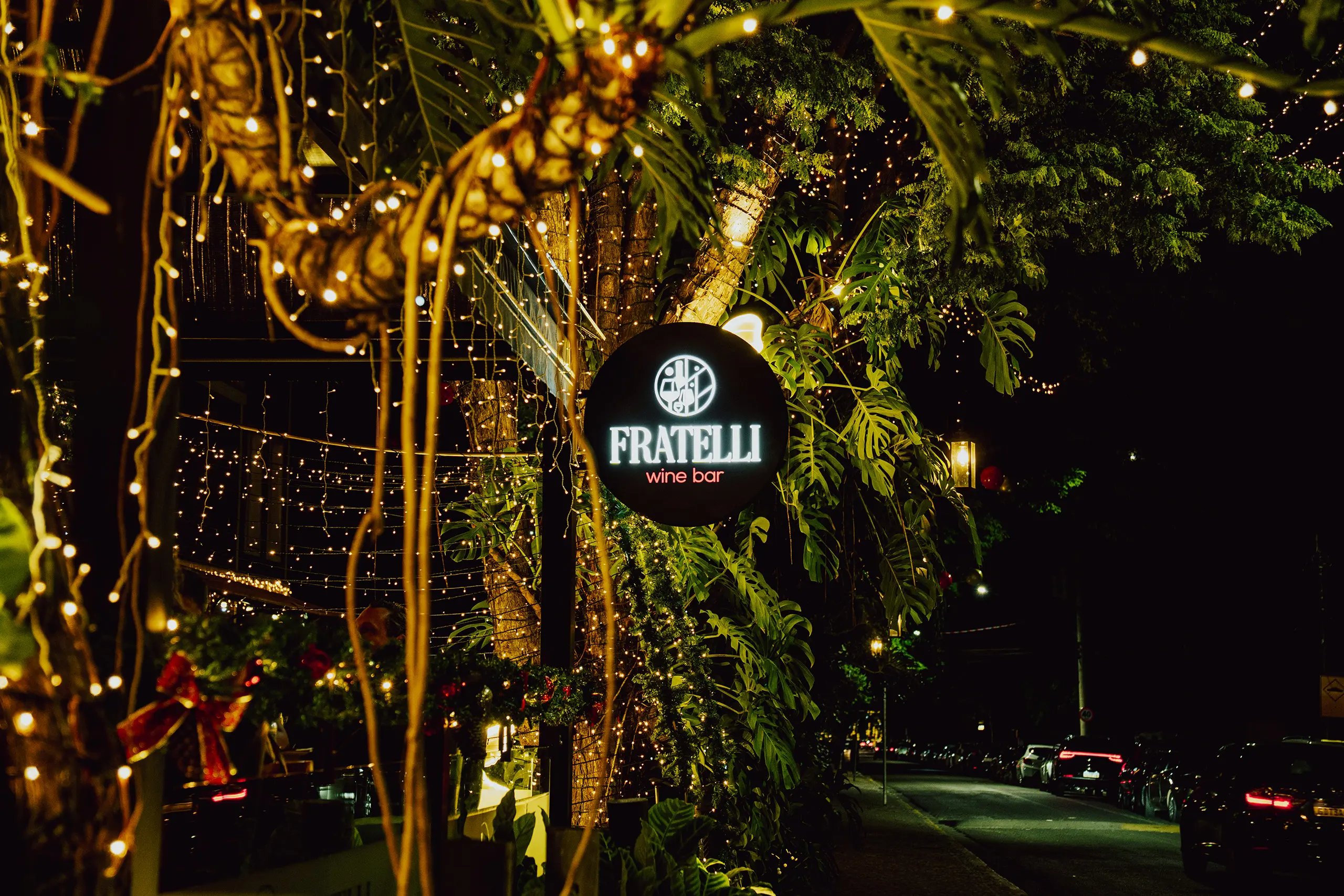 Placa iluminada do Fratelli Wine Bar entre árvores e luzes decorativas à noite em Holambra
