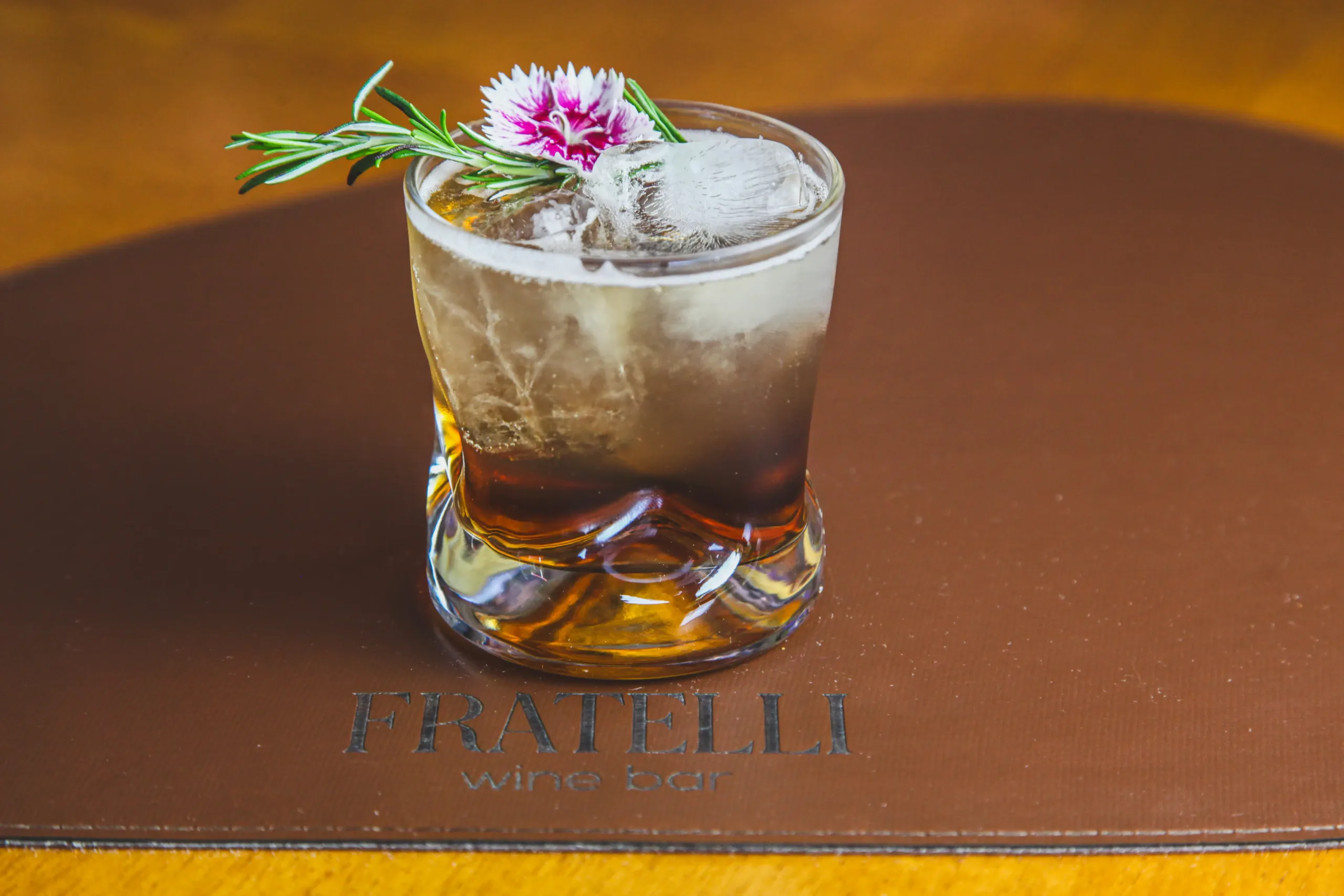 Drink com base alcoólica escura, gelo, alecrim e flor comestível servido no Fratelli Wine Bar em Holambra.